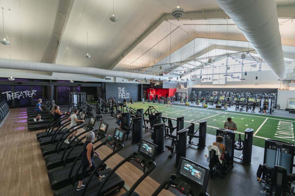 A light filled fitness floor, with people using cardio machines, an open turf area, and weights area.