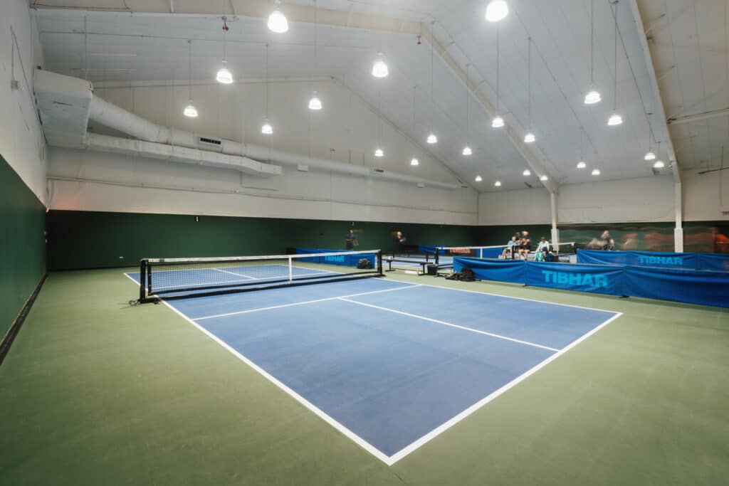 People play pickleball on one of three pickleball courts that sit under a tall, white roof. Large lights hang from the ceiling.